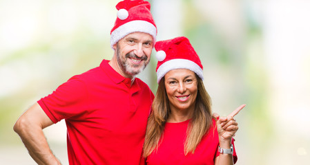 Middle age hispanic couple wearing christmas hat over isolated background with a big smile on face, pointing with hand and finger to the side looking at the camera.