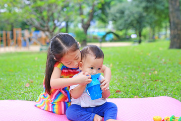 Adorable Asian sister sitting on pink mattress mat take care her little brother to drinking water from Baby sippy cup with straw in the park garden.