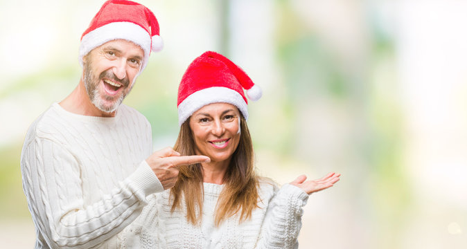 Middle age hispanic couple wearing christmas hat over isolated background amazed and smiling to the camera while presenting with hand and pointing with finger.