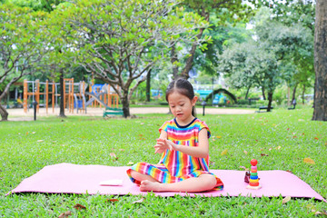 Adorable little Asian child girl sitting on pink mattress and applying body lotion in the summer garden.