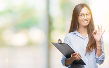 Obraz premium Young asian business woman holding clipboard over isolated background doing ok sign with fingers, excellent symbol
