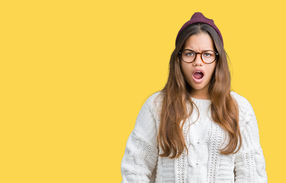 Young beautiful brunette hipster woman wearing glasses and winter hat over isolated background In shock face, looking skeptical and sarcastic, surprised with open mouth
