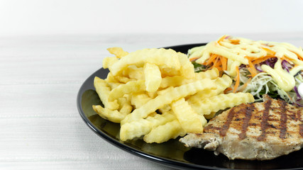 Pork steak in a dish on a white wood floor table garnish spicy sauce with French fries, green salad, purple cauliflower, Front view.