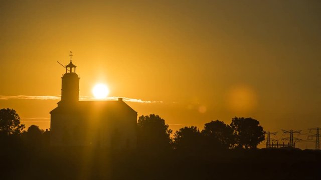 Time lapse of the sunset behind the silhouette of a little white church standing in the country side  with trees and power lines on the horizon