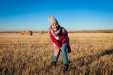 Naklejka na ściany i meble Young attractive lady in gray knitting hat and pink coat posing around haystick. Smiling beautiful hipster happy woman in field, wearing stylish clothes. Autumn trend, urban stydent style.