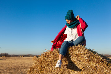 Naklejka na ściany i meble Portrait of a beautiful young model in blue knitted hat and warm clothes have fun, sitting on haystacks in sunny autumn day . Autumn warm photo. Woman smiling and look away, joyful cheerful mood.