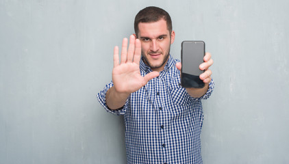 Young caucasian man over grey grunge wall showing blank screen of smartphone with open hand doing stop sign with serious and confident expression, defense gesture