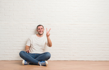 Young caucasian man sitting on the floor over white brick wall smiling looking to the camera showing fingers doing victory sign. Number two.