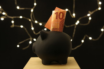 Czarna świnka skarbonka z banknotem 10 euro - ciemne tło z lampkami - obrazy, fototapety, plakaty