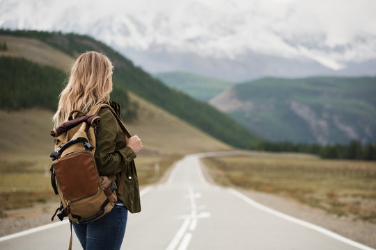 A woman with a backpack and a road stretching into the distance against the backdrop of mountains