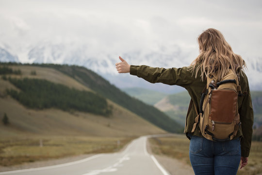 A woman with a backpack and a road stretching into the distance against the backdrop of mountains - Powered by Adobe