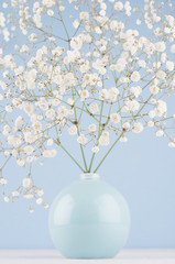 Spring branch with small white flowers in blue sphere vase on soft light blue modern wall and white table, closeup.