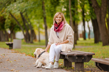 A young woman sits on a bench in the park, and her Labrador walks nearby.