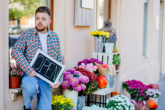 Young florist man holding sign open outdoor of flower shop near bouquets, autumn decoration, pumpkins. Concept of hospitable private business floristic art.