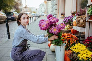 Friendly positive woman florist in apron taking care of chrysanthemums flowers outdoor on morning city street on background Concept of hospitable private business floristic art.