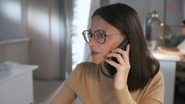 Close-up of attractive brunette business lady in eyeglasses using computer and having phone conversation while working late in the office