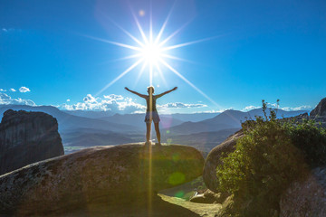 Fototapeta na wymiar Girl traveler with open arms at the sun, on a rock on sunset in Meteora landscape in Thessaly, Greece. Panoramic view from best view point Psaropetra. Tourism in Europe.