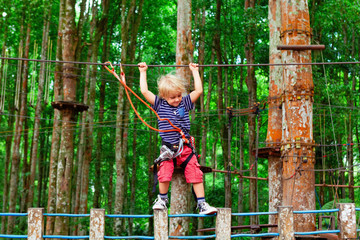 Brave young child in safety harness climb high on tree tops, pass hanging bridge obstacle in adventure rope park. Outdoor activity, active lifestyle in summer scout camp on family holidays with kids