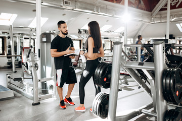 Fototapeta na wymiar Dark-haired athletic girl dressed in black sports top and tights with towel on her neck talks with fitness coach in the gym