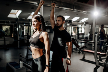 Fototapeta na wymiar Dark-haired athletic girl dressed in black sport clothes and fitness coach are doing warm up in the gym