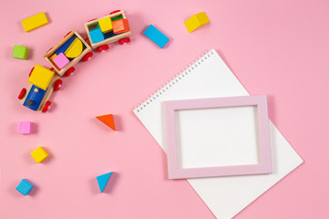 Wooden toy train, colorful cubes and white open notebook anf frame on pink background