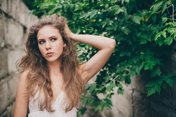 Beautiful mad girl with curly natural hair in white dress near green tree leaves. Summer beauty portrait. Crazy lady in nature outdoors. Alone moody woman with pensive sight in long tunnel in forest.