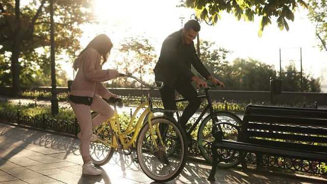 Beautiful young couple riding their bikes in the empty city park. Stop and sit on the bench and embracing. Loving couple. Sun shines on the background. Accelerating footage