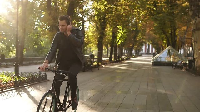 Young caucasian man with headphones riding bicycle in the city park on the trekking black bike. Riding and feeling good while listening to the favourite music. Sun shines on the background