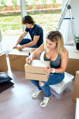 Sitting woman labeling moving boxes while her husband prepares boxes