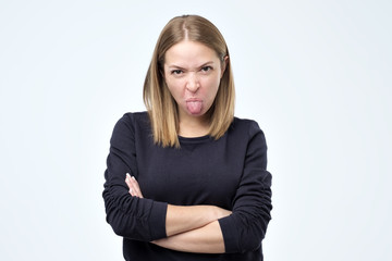 Dissatisfied female model frowns face, has disgusting expression, shows tongue, expresses disgust, irritated with somebody, rejects do something.