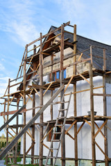 Fototapeta na wymiar Wooden scaffolding next to white unfinished residential house with grey shingles roof on process of facade insulation works with polyurethane styrofoam. House improvement process nd DIY concept.