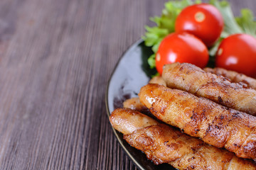 Appetizing and colorful sausages wrapped in bacon and grilled next to tomatoes and lettuce are on a wooden table on the right