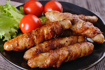Appetizing and colorful sausages wrapped in bacon and grilled next to tomatoes and lettuce are on a wooden table