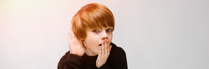 Charming teenager with blond hair and dark eyes. The boy closes his mouth with one hand, brings his other hand to his ear