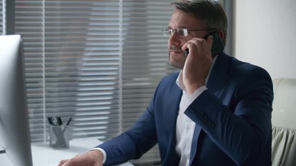 Caucasian middle aged businessman in eyeglasses and formal suit using computer and talking on mobile phone at office desk in the evening - Powered by Adobe