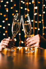 cropped image of couple clinking with glasses of champagne on garland light background, christmas concept