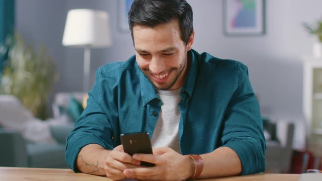 Handsome Smiling Man Uses Smartphone while Sitting at the Desk of His Living Room. Guy Browsing in Internet, Using Social Networks, Relaxing at Home.