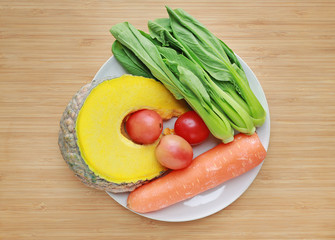 Variety of fresh vegetables on white plate against wooden board background.