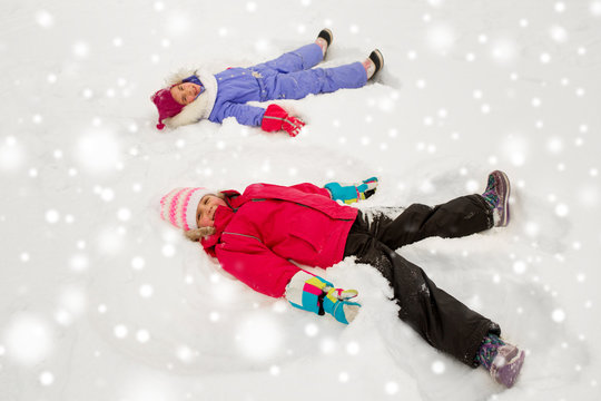 childhood, leisure and season concept - happy little girls in winter clothes making snow angels outdoors