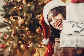 Merry Christmas. happy girl in santa hat holding many gift boxes at golden beautiful christmas tree...