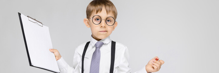 A charming boy in a white shirt, suspenders, a tie and light jeans stands on a gray background. The...