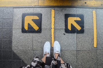 Top view of woman feet standing over yellow arrow sign on subway platform at the train station.