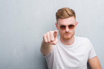 Young redhead man over grey grunge wall wearing retro sunglasses pointing with finger to the camera...