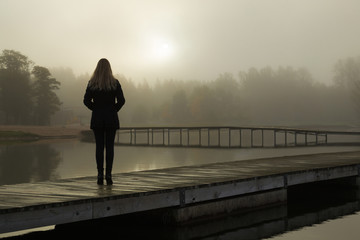 Young woman standing alone on lake footbridge and staring at sunrise in gray, cloudy sky. Mist over...