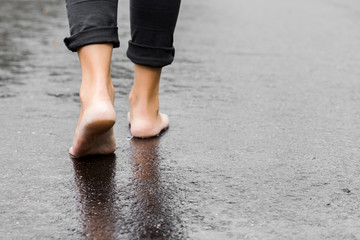 Young woman's barefoot walking on the wet, dark black asphalt after warm rain. Cloudy day in...