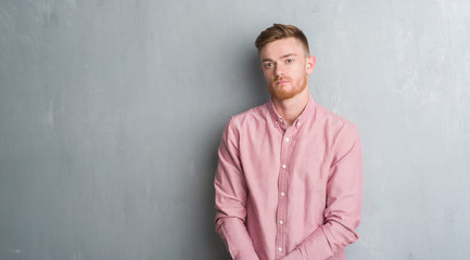 Fototapeta premium Young redhead man over grey grunge wall wearing pink shirt with serious expression on face. Simple and natural looking at the camera.