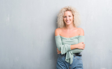 Young blonde woman with curly hair over grunge grey background happy face smiling with crossed arms looking at the camera. Positive person.