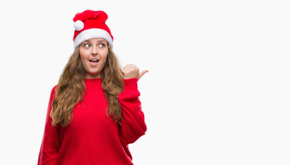 Young blonde woman wearing santa claus hat pointing and showing with thumb up to the side with happy face smiling