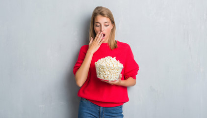Beautiful young woman over grunge grey wall eating pop corn cover mouth with hand shocked with shame for mistake, expression of fear, scared in silence, secret concept