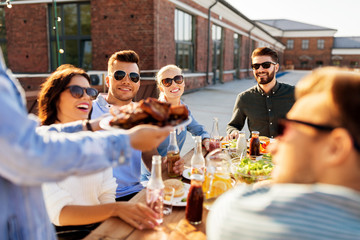 leisure and people concept - happy party host offering meat to his friends at barbecue party on rooftop in summer